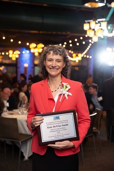 person standing interior in a salmon blazer smiling at camera holding award plaque
